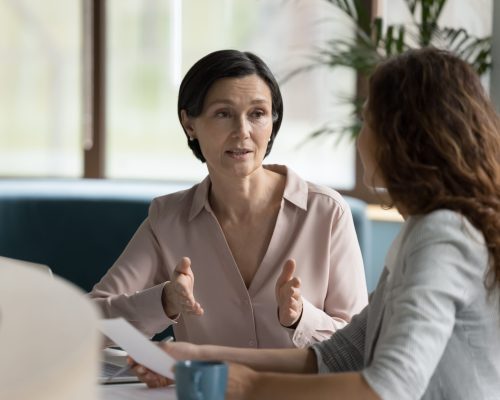 Confident mature business professional woman talking to younger female colleague at office table, speaking, gesturing, teaching, explaining work tasks. Elder mentor training intern